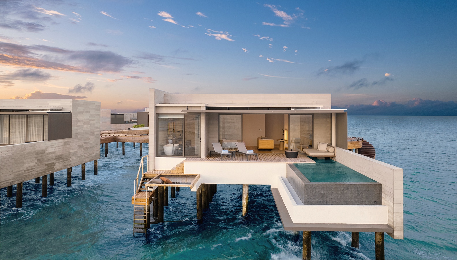 An overwater pool villa at dusk. The deck extends directly above the water, pool flush with the lagoon edge.