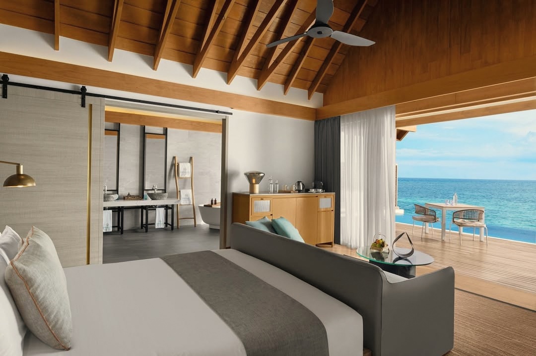 The interior of an overwater villa. Clean lines, lagoon light through full-height glass, the water visible from the bed.