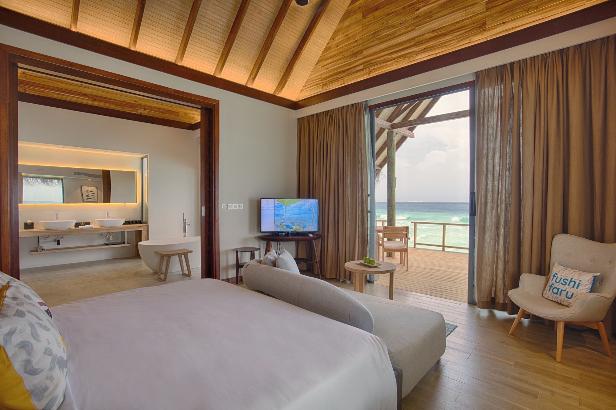 The interior of a sunset-facing water villa. West orientation, warm evening light, the lagoon visible from the bed.