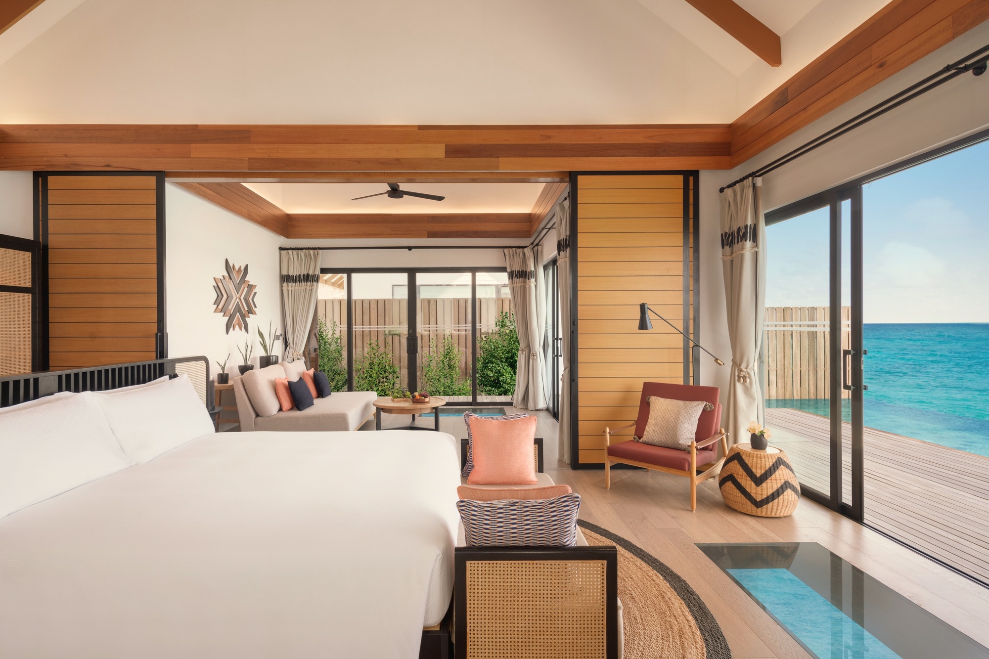 The villa interior looking toward the glass floor panel. Natural materials, open plan, the lagoon visible beneath the room.