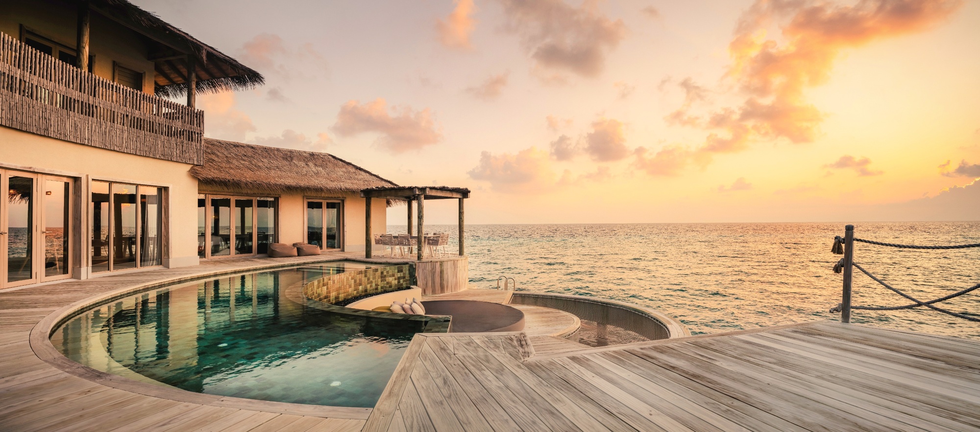 An overwater pool villa at dusk. The deck extends directly above the lagoon, with the reef visible below at low tide.
