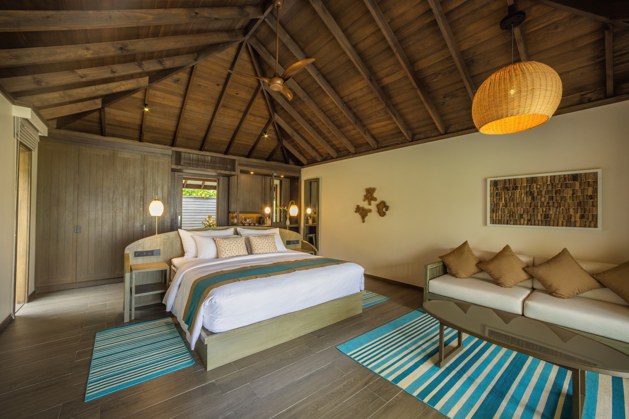 The interior of a beach villa. White sandy beach visible through the full-length glass, with the lagoon beyond.