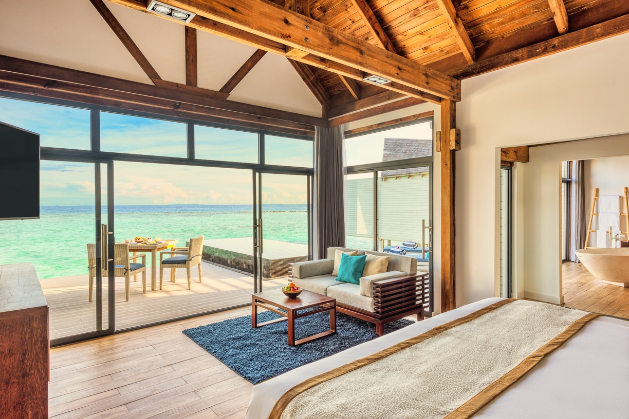 The interior of an overwater villa looking out toward the horizon. Full-length glazing and a private pool at the threshold between room and ocean.