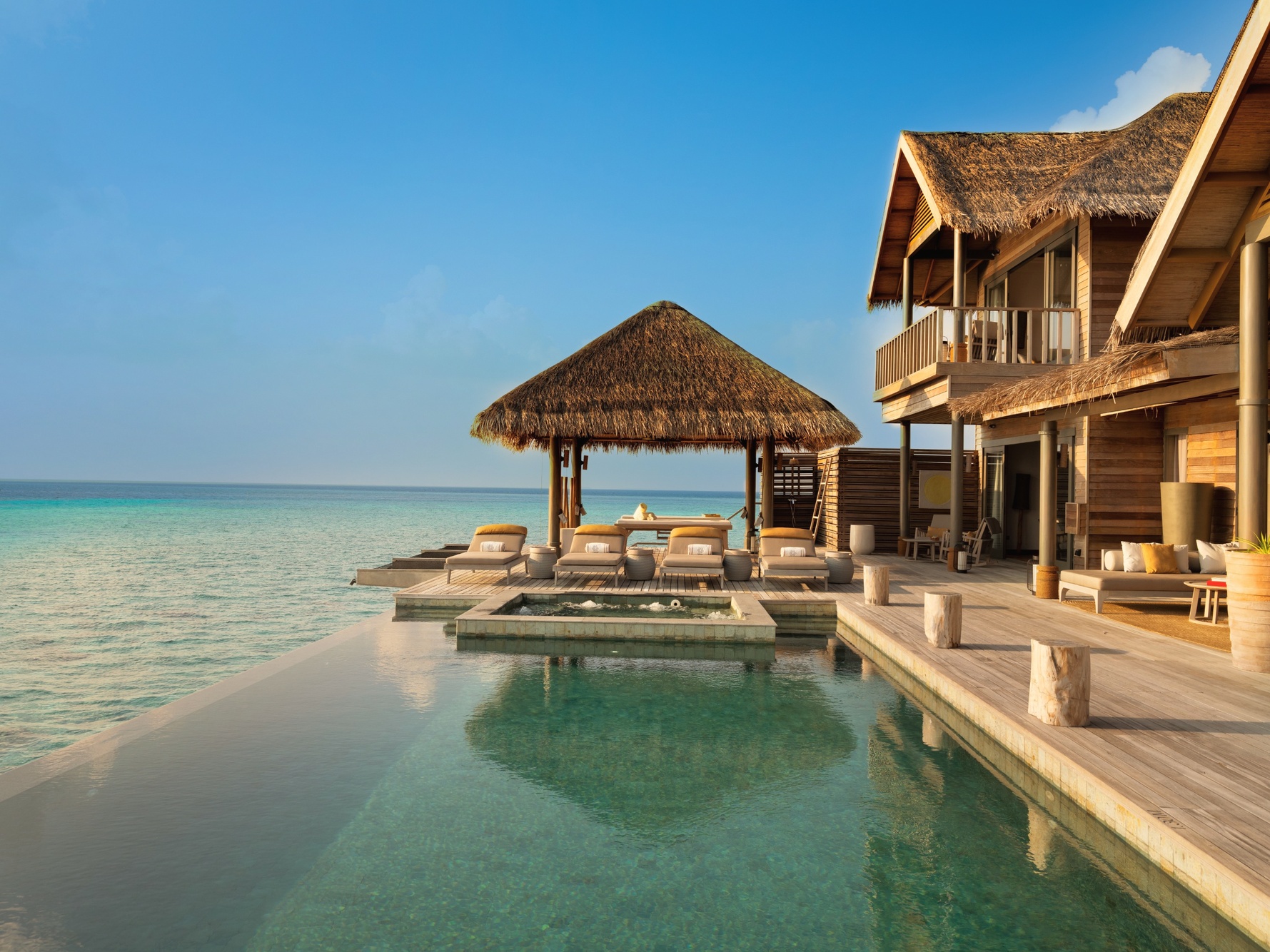 An overwater pool villa at dusk. The deck extends directly over the lagoon, with the reef visible in the shallows below.